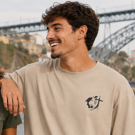 Homem sorridente com cabelo encaracolado usando camiseta bege com estampa pequena em preto de rosto estilizado e cruz, ao ar livre com ponte ao fundo.
